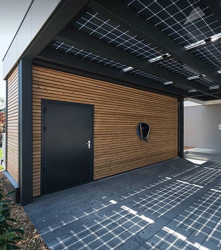 Modernes Holzgebäude mit einer dunklen Tür, einem Dach mit Sonnenkollektoren und einem raffinierten Schattenwurf auf dem Gehweg bei hellem Tageslicht.